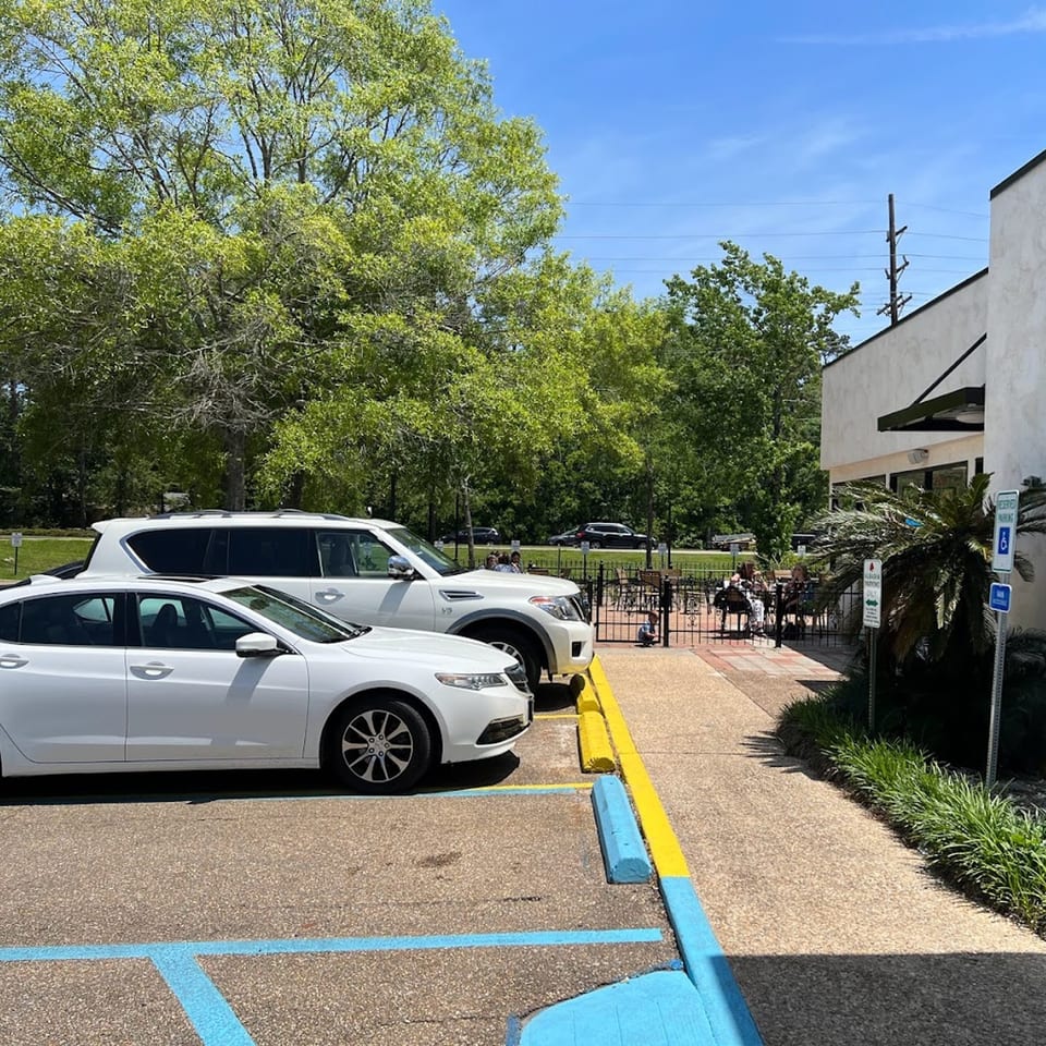 Albasha Express restaurant exterior with parked cars, outdoor patio seating, and lush green trees under a blue sky.