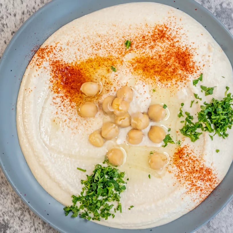 Creamy hummus topped with whole chickpeas, olive oil, paprika, and fresh parsley on a blue plate.